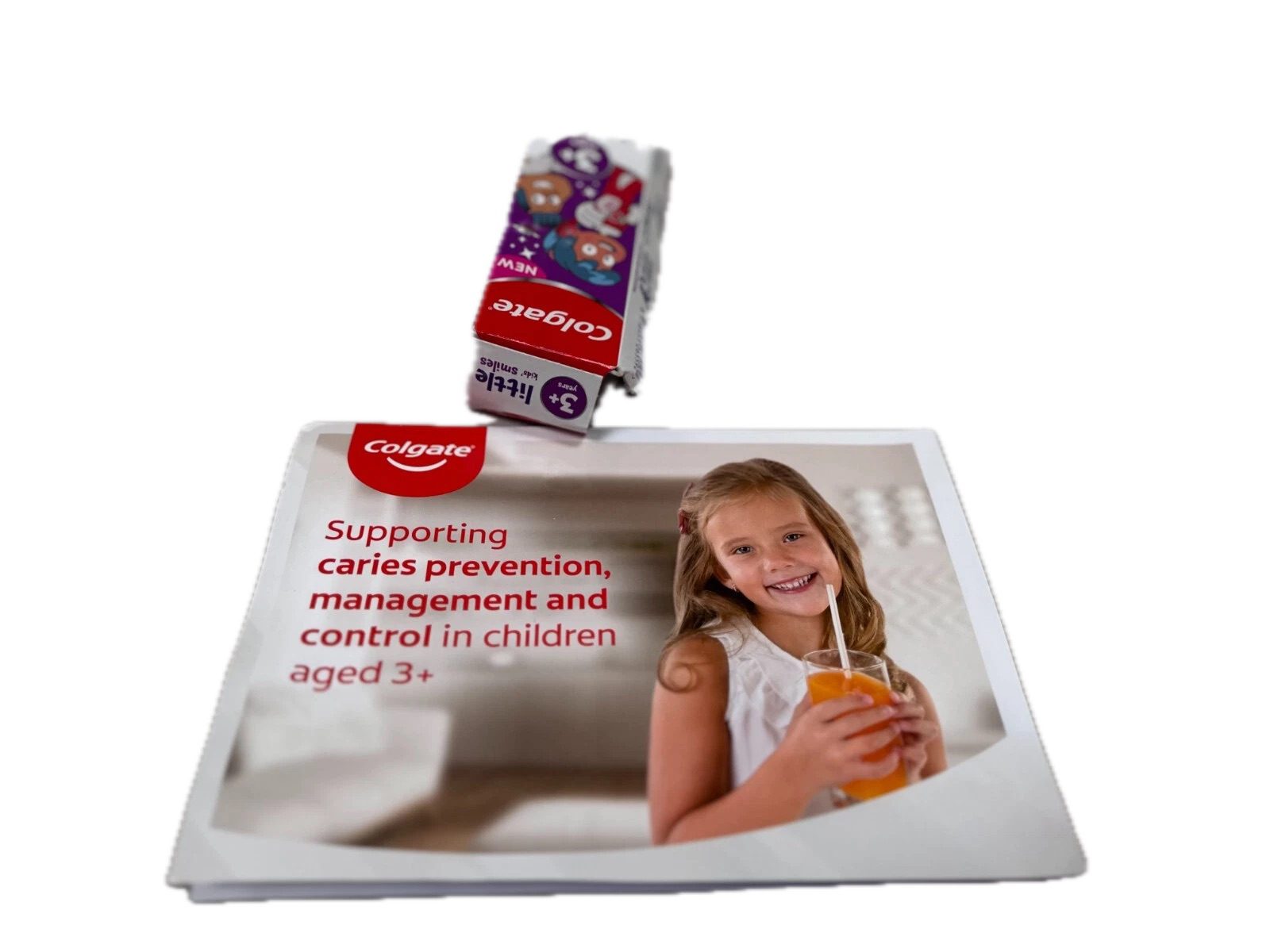Two Colgate Kids’ Toothpaste boxes (3+ and 6+) shown side by side on a white table, with a dental care leaflet featuring a smiling child holding a juice glass.
